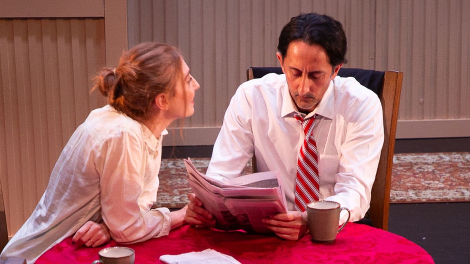 Two actors at a breakfast table on stage: a woman leans in toward a man reading the newspaper